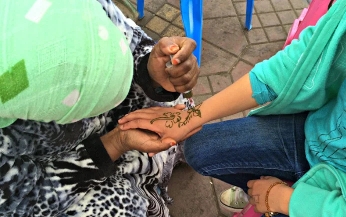 A woman applies a complex henna design to a hand at an outdoor market stall.