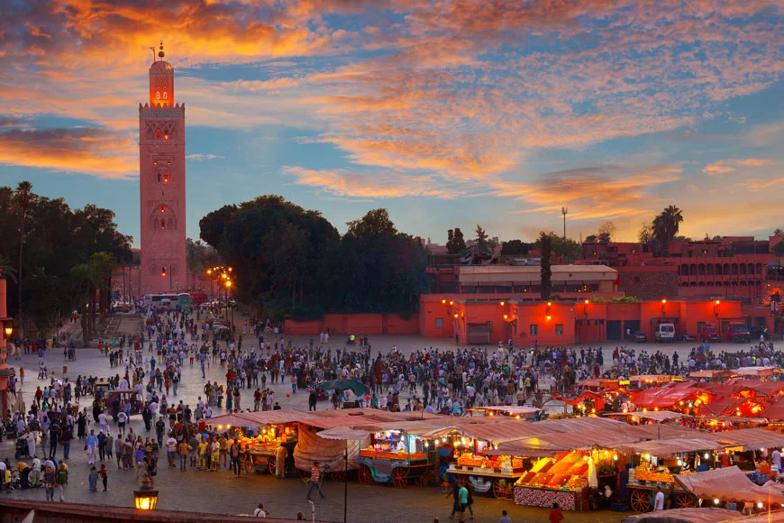 Crowds gather in a bustling square with food stalls under a sunset sky.