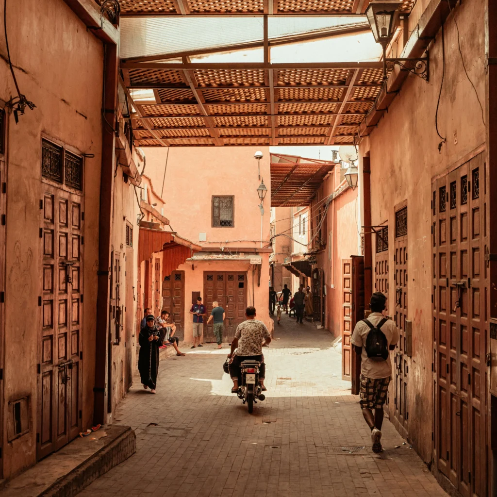 A narrow sunlit alleyway in the Marrakech Medina with reddish-brown walls.