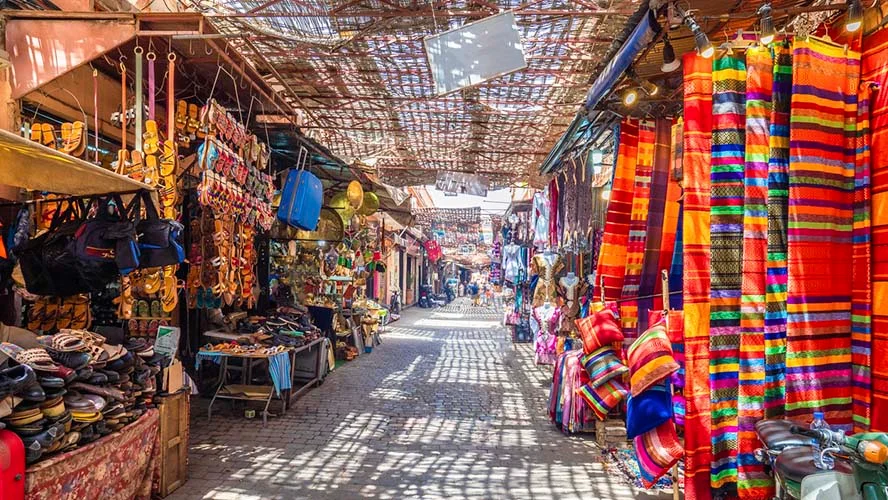 Colorful striped textiles and leather goods line a narrow, shaded market.