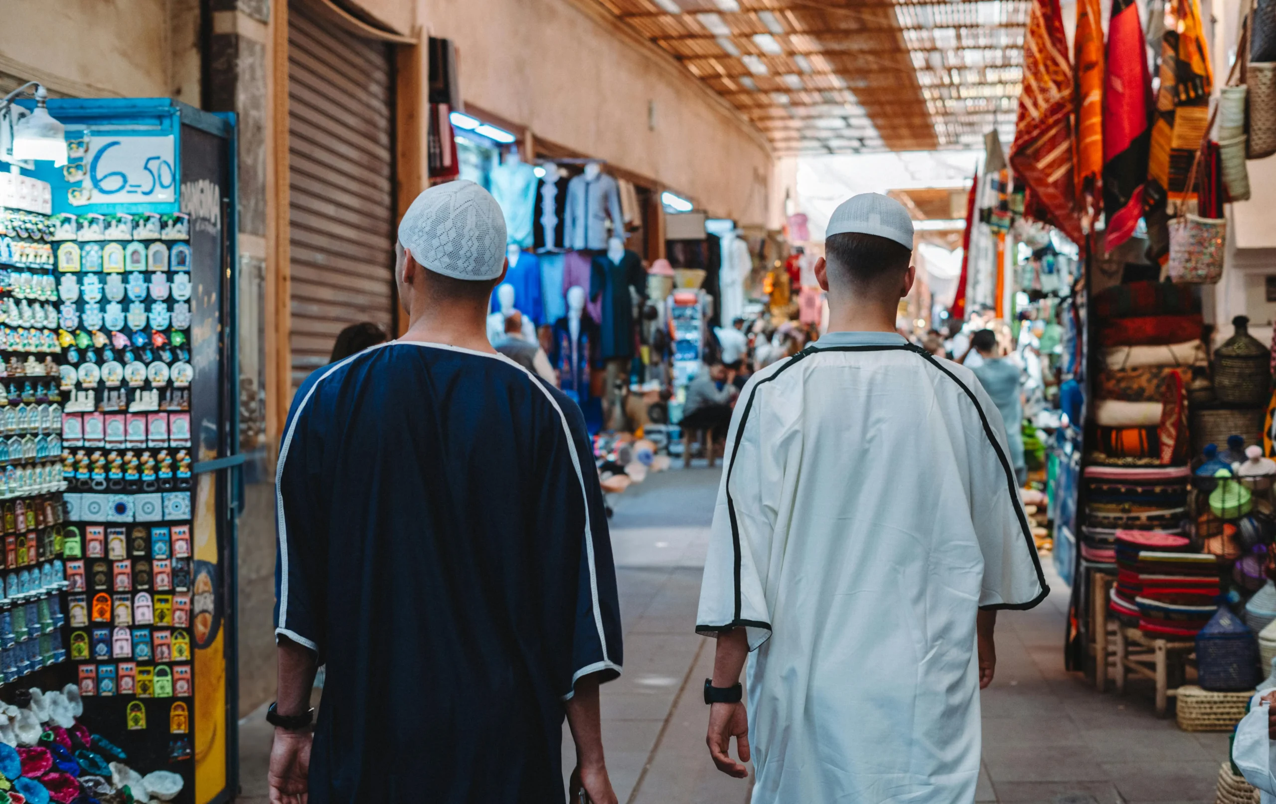 Rear view of two men wearing traditional djellabas and prayer caps walking through a covered market alley in Morocco, surrounded by stalls of textiles and souvenirs.
