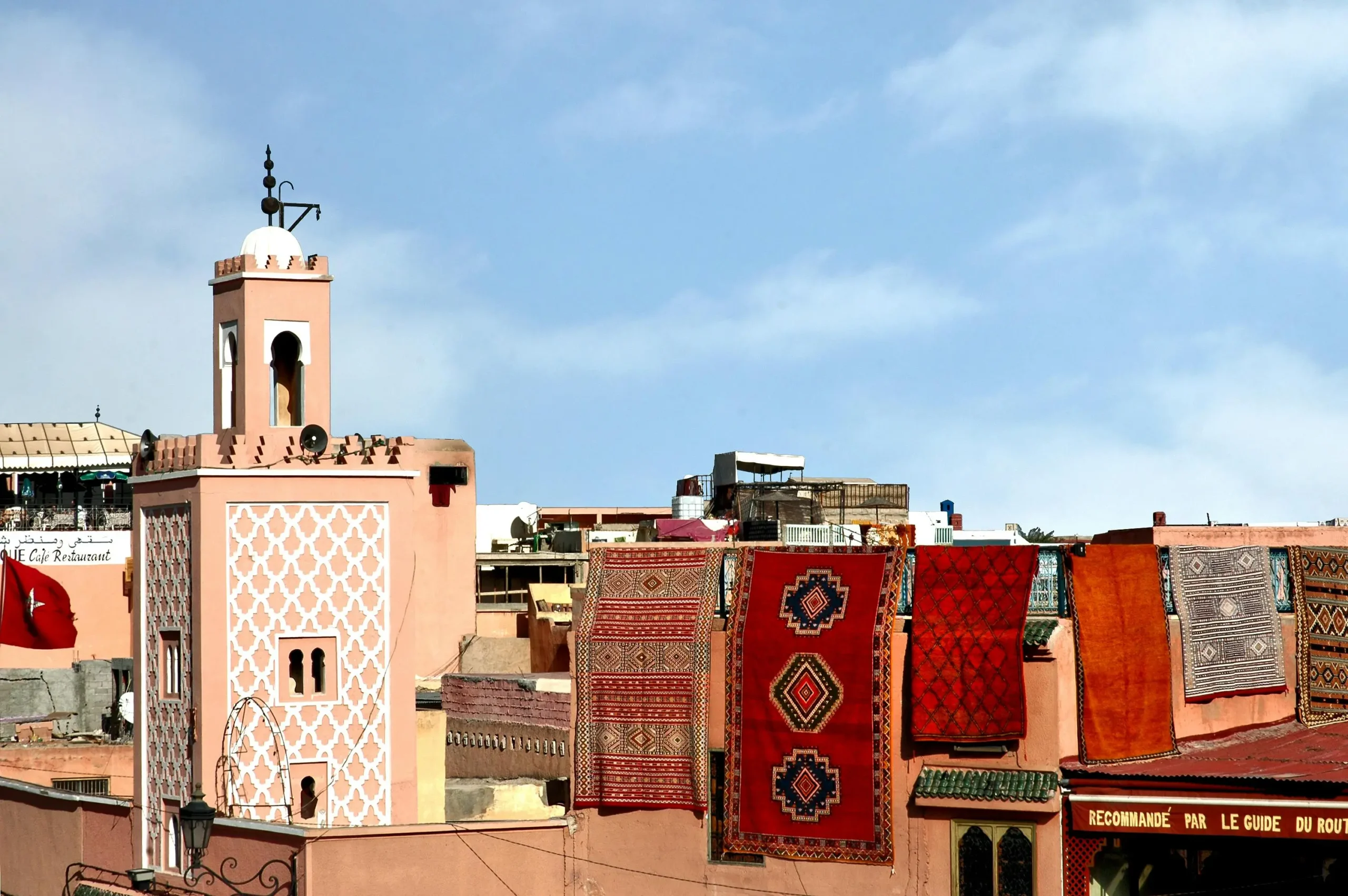 A scenic view of Marrakech rooftops featuring a salmon-colored minaret with white geometric patterns, with several traditional Berber rugs hanging on a wall to dry under a blue sky.