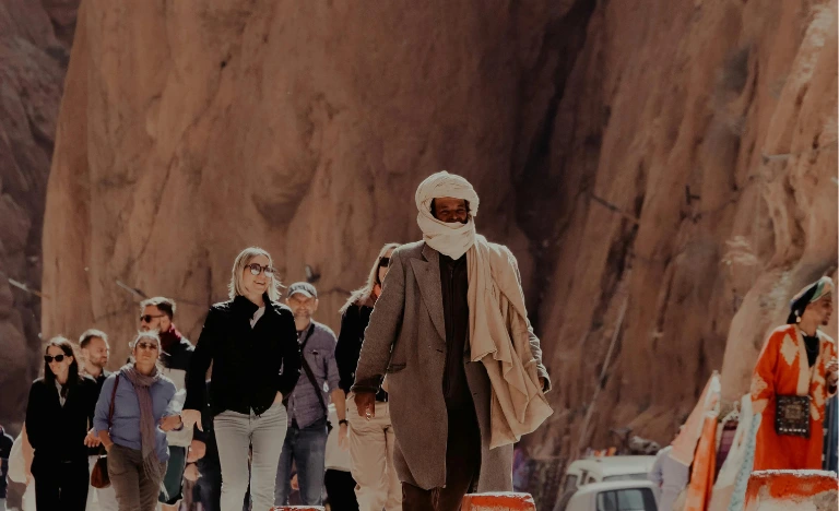 A guide in a turban leads a group of tourists through a rocky desert canyon.