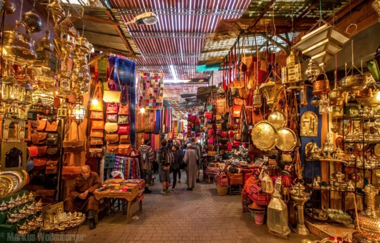 A narrow market aisle packed with hanging brass lamps and leather goods.