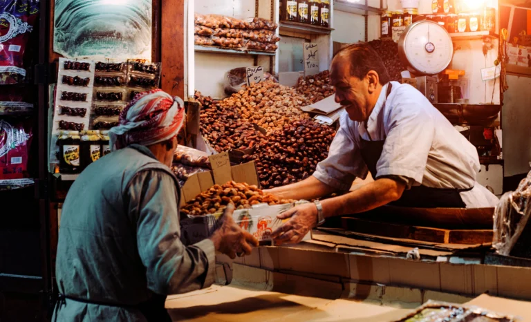 A vendor hands a box of fresh dates to a customer in a busy market stall.