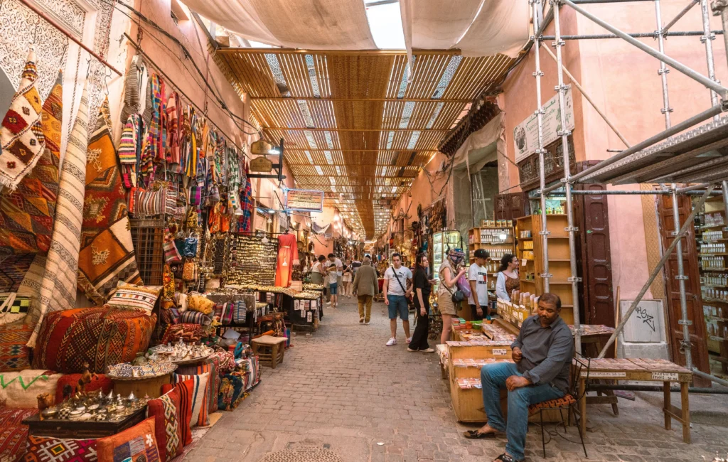 Tourists walk through a busy outdoor market street lined with textiles.