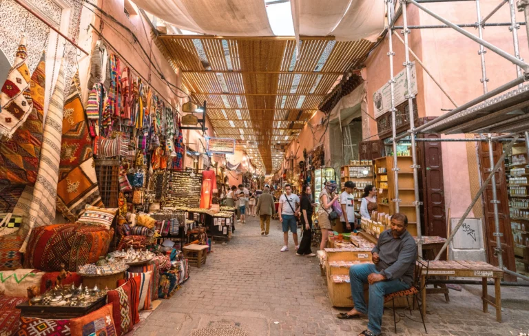 Tourists walk through a busy outdoor market street lined with textiles.
