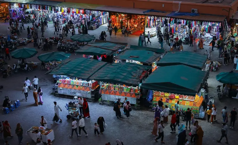 Brightly lit fruit stalls and crowds at Jemaa el-Fna market at night.