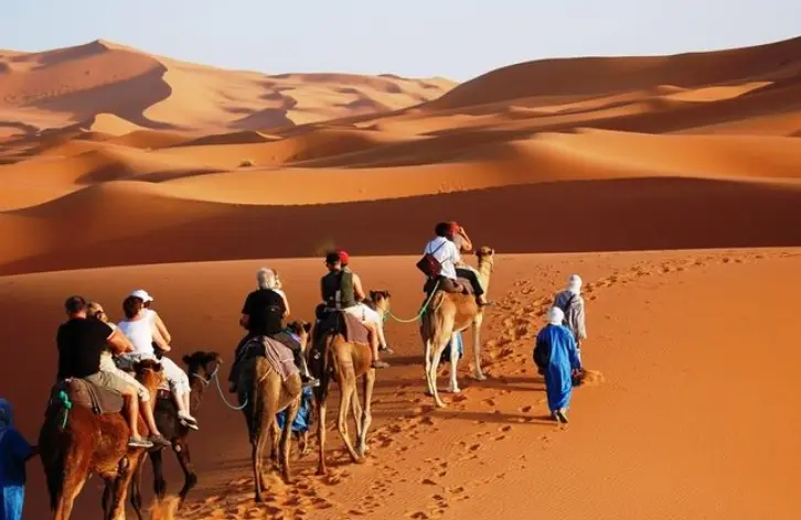 Tourists riding camels across golden desert sand dunes under a clear sky.