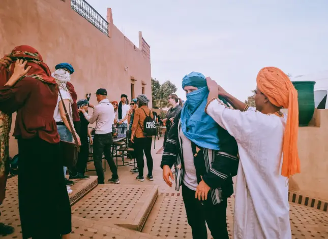 Local man helping a traveler wrap a blue turban in a traditional setting.