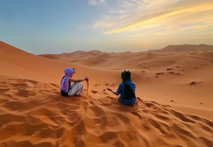 Two people sitting on a sandy dune chatting during a golden sunset.
