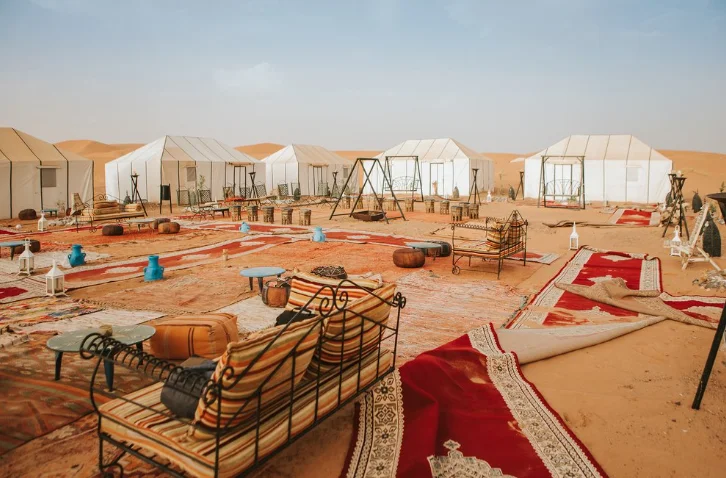 Spacious desert camp with traditional carpets, seating, and white tents.
