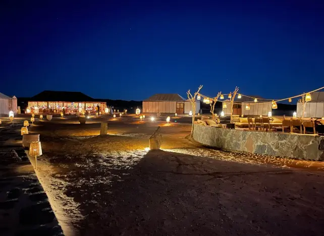 Illuminated desert camp with warm lanterns under a deep blue night sky.