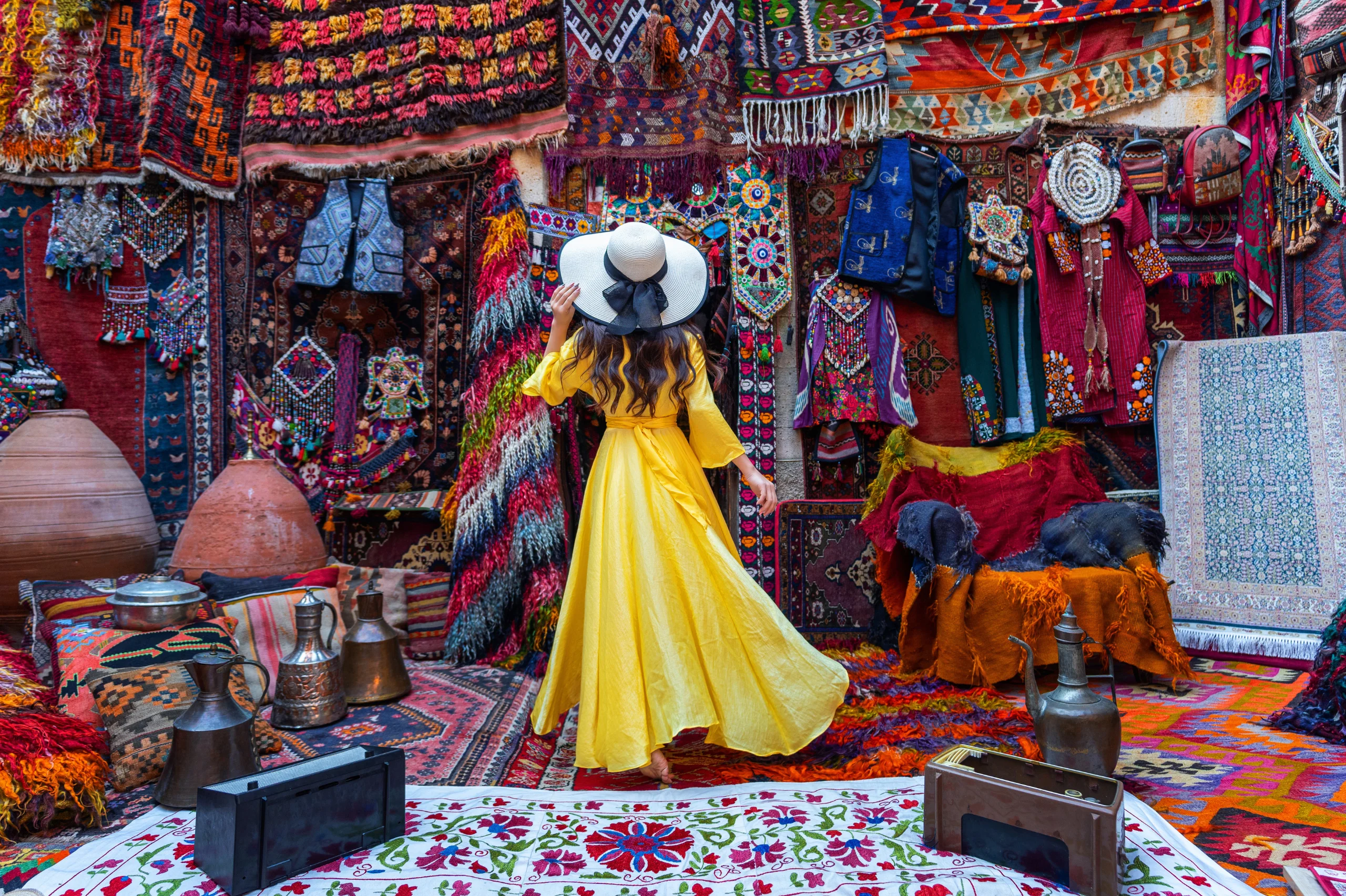 A woman in a yellow dress looks at many vibrant hanging rugs.